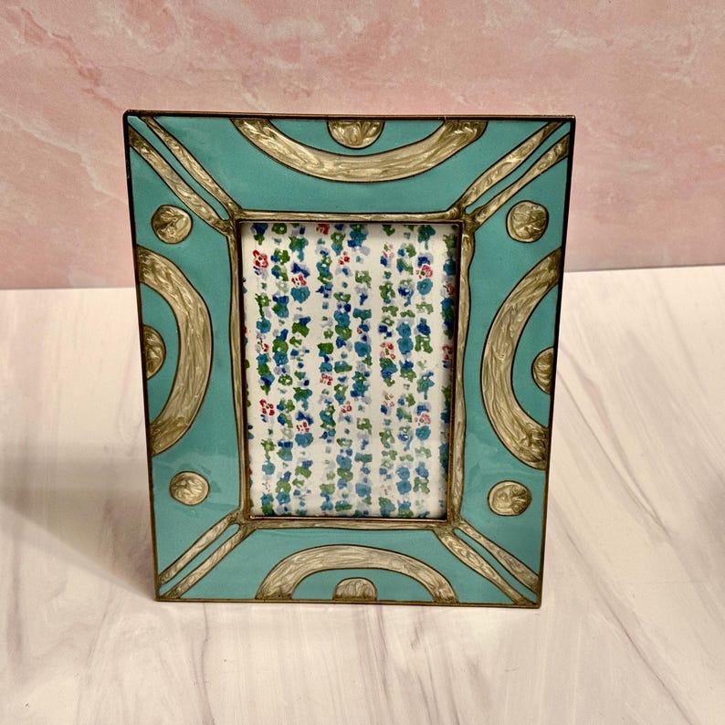 Puede incluir: Un marco de fotos rectangular con acabado esmaltado turquesa y detalles dorados. El marco tiene un inserto con estampado floral. El marco mide aproximadamente 15 cm de alto y 10 cm de ancho.