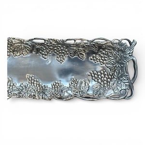 May include: Silver-toned rectangular serving tray with grape and leaf design. The tray features detailed relief work of grape clusters and leaves around the edges and handles. The center is smooth and reflective.