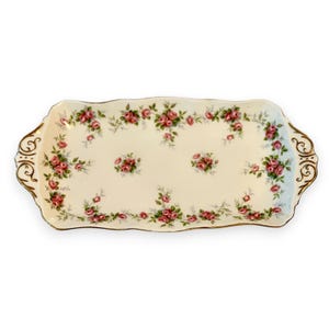May include: A rectangular porcelain serving tray with a cream-colored base, featuring a repeating pattern of pink roses and green leaves. The tray has scalloped edges and gold-trimmed handles on both sides with ornate, swirling gold detailing.