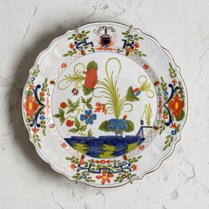 May include: White ceramic plate with a scalloped edge, featuring a central floral design in red, blue, and green. The plate has a gold rim and is held by a metal stand. The plate has the word "Giardino" written on it.
