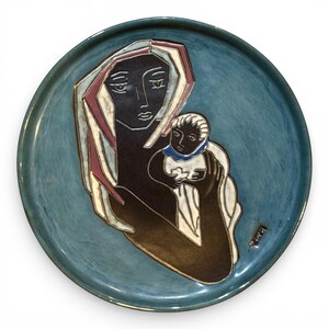 May include: A ceramic plate with a teal blue rim and background. The plate features a stylized depiction of a mother and child in dark brown, white, and red. The mother figure holds the child, both with serene expressions.