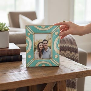 Puede incluir: Marco de fotos rectangular turquesa y dorado, con una foto de una pareja sonriente. El marco est&aacute; sobre una mesa de madera. Una mano sostiene el marco.