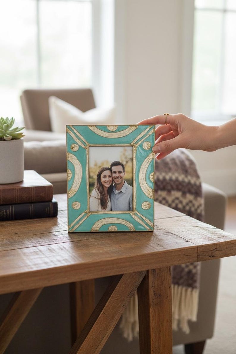Puede incluir: Marco de fotos rectangular turquesa y dorado, con una foto de una pareja sonriente. El marco est&aacute; sobre una mesa de madera. Una mano sostiene el marco.