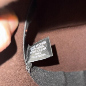 May include: Close-up of a brown fedora hat with a black band. A tag inside the hat reads "MADE IN CHINA 100% WOOL SPOT CLEAN ONLY RN#00834". The hat is made of wool.