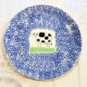 May include: A blue and white ceramic plate with a black and white cow illustration in the center. The plate has a speckled blue glaze.