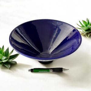 May include: A deep blue ceramic bowl with a glossy finish. The bowl has a wide, shallow design and sits on a black base. A pen is placed near the bowl for scale, and two succulents are in the background.