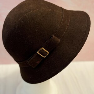 May include: A dark brown cloche hat with a small brim and a decorative band. The band features a gold-colored buckle. The hat has a rounded crown and is made of a soft, felt-like material. The hat is a vintage style.