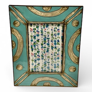 Puede incluir: Un marco de fotos decorativo con fondo turquesa y detalles dorados. El marco tiene una abertura rectangular con un estampado floral visible en el interior. El dise&ntilde;o del marco incluye l&iacute;neas curvas y detalles circulares.