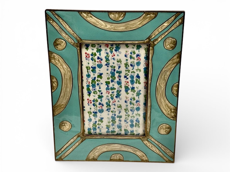 Puede incluir: Un marco de fotos decorativo con fondo turquesa y detalles dorados. El marco tiene una abertura rectangular con un estampado floral visible en el interior. El dise&ntilde;o del marco incluye l&iacute;neas curvas y detalles circulares.
