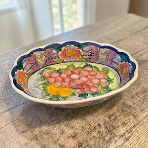 May include: Oval ceramic serving bowl with a scalloped edge. The bowl features a hand-painted design of pink grapes, yellow fruit, and green leaves. The rim is decorated with floral and scroll patterns in pink, blue, and yellow.