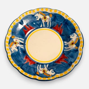 May include: A round ceramic plate with a scalloped edge featuring a central cream-colored circle bordered by a yellow ring. The outer rim displays a pattern of blue brushstrokes, red accents, and a border of yellow dots against a white background.