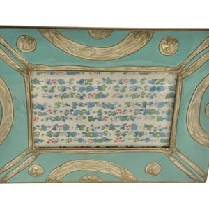 Puede incluir: Marco de fotos rectangular con acabado esmaltado turquesa y detalles plateados. El marco tiene un dise&ntilde;o decorativo con l&iacute;neas curvas y c&iacute;rculos. El interior contiene una impresi&oacute;n con motivos florales. Un art&iacute;culo decorativo para la decoraci&oacute;n del hogar.
