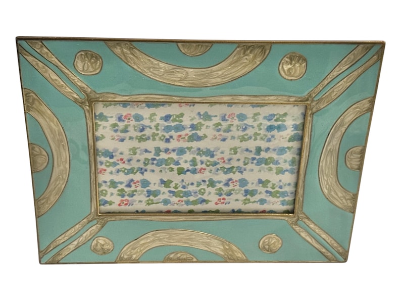 Puede incluir: Marco de fotos rectangular con acabado esmaltado turquesa y detalles plateados. El marco tiene un dise&ntilde;o decorativo con l&iacute;neas curvas y c&iacute;rculos. El interior contiene una impresi&oacute;n con motivos florales. Un art&iacute;culo decorativo para la decoraci&oacute;n del hogar.