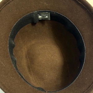May include: A brown fedora hat with a black inner band. The hat's interior is a lighter shade of brown, with a stitched seam running down the center. The label reads "AUGUST 16".