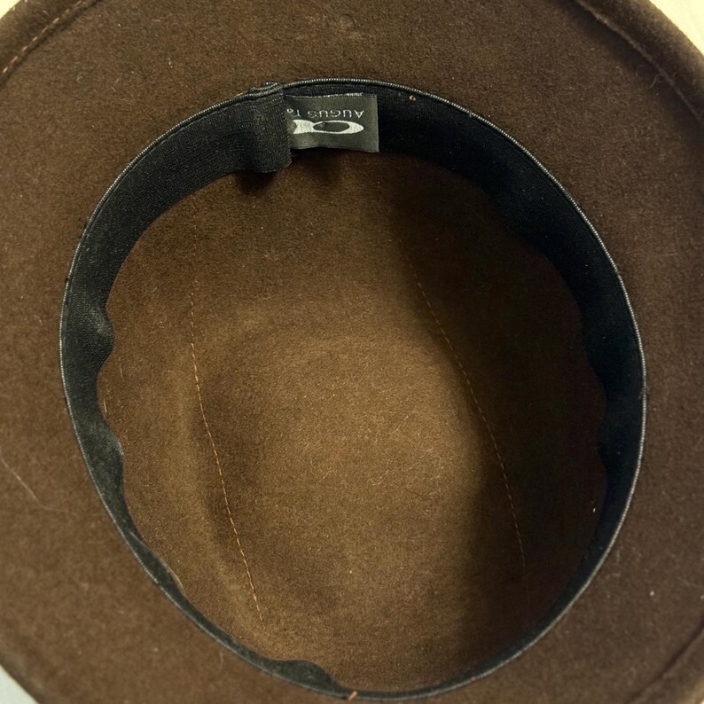 May include: A brown fedora hat with a black inner band. The hat's interior is a lighter shade of brown, with a stitched seam running down the center. The label reads "AUGUST 16".