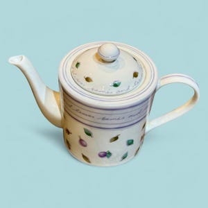 May include: A white ceramic teapot with a lid, spout, and handle. The teapot is decorated with purple stripes, green leaves, and purple berries. The lid has a small knob and the words "leaves, berries and leaves" are written on the teapot.