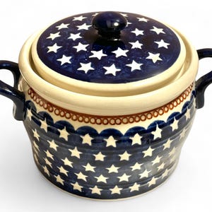 May include: Ceramic casserole dish with a lid, featuring a navy blue and white star pattern. The dish has two handles and a dark blue knob on the lid. The rim is cream-colored with a brown and white scalloped design.