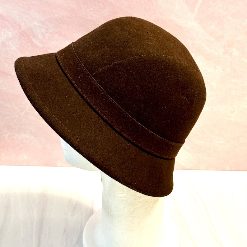 May include: A brown cloche hat with a rounded crown and a small brim. The hat features a band around the crown. The hat is displayed on a white mannequin head against a pink background.