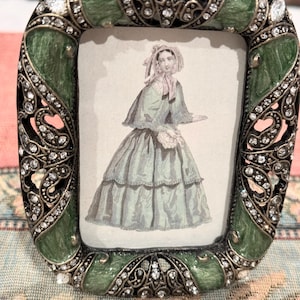 May include: A small, ornate picture frame with a green enamel finish and a decorative design. The frame is adorned with rhinestones and features a cutout design. The frame is holding a vintage print of a woman in a dress.