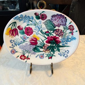 May include: An oval ceramic serving platter featuring a white background decorated with a vibrant botanical print of various flowers including red tulips, purple hydrangeas, blue delphiniums, and green foliage with a thin pink rim.