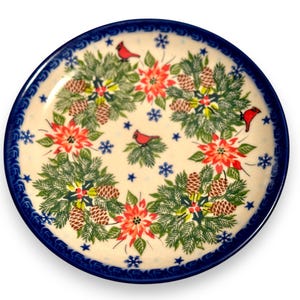 May include: A round ceramic plate with a cream-colored base and a dark blue rim. The surface features a repeating pattern of green pine branches, brown pinecones, red poinsettia flowers, small red cardinal birds, and blue snowflakes.