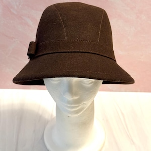 May include: A brown cloche hat with a small bow on the side. The hat has a rounded crown and a slightly downward-sloping brim. The hat is displayed on a white mannequin head against a pink background.