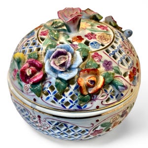 May include: A decorative, round porcelain container with a lid, adorned with colorful, three-dimensional roses and floral accents. The container features a lattice design and gold trim, creating an elegant, vintage aesthetic.
