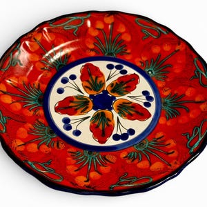 May include: A decorative ceramic plate with a vibrant red background and a scalloped edge. The plate features a central design with a white circle, blue accents, and stylized floral patterns in orange, red, and green.