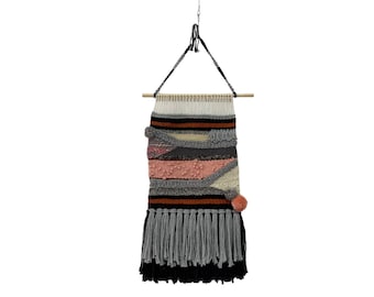 May include: A woven wall hanging with a geometric pattern in shades of gray, pink, orange, and black. The hanging has a fringe border and two small pom-poms.