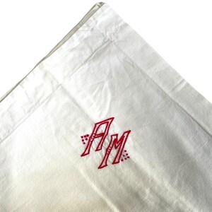 Peut inclure: Tissu de lin blanc avec des initiales brodées en rouge "AM" dans une police décorative.