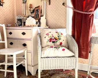 Könnte beinhalten: Ein weißer Korbstuhl mit einem floral bestickten Kissen steht auf einem Teppich mit Blumenmuster in einem Raum mit einer weißen Kommode und einem Spiegel mit einer Muschel darauf. Rote Samtvorhänge sind im Hintergrund.