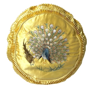 Op de afbeelding: Een ronde, goudgele decoratieve kussen met een geplooid rand. De kussen heeft een geborduurde pauw met een blauw lichaam en een waaierstaart van groene, witte en blauwe veren. Er is ook een andere vogel geborduurd.