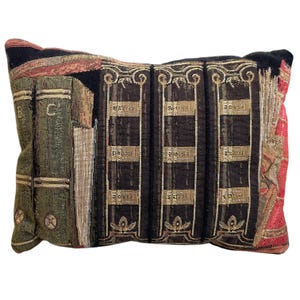 May include: A decorative pillow with a tapestry design featuring a bookshelf with books and a repeating pattern of black and gold squares. The pillow is rectangular and has a red, gold, and green color scheme.