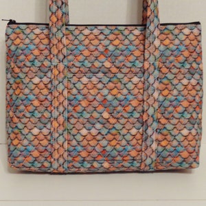 Könnte beinhalten: Eine gesteppte Tragetasche mit einem Fischschuppenmuster in Orange-, Blau- und Pfirsichtönen. Die Tasche hat eine rechteckige Form, eine Vordertasche, einen schwarzen Reißverschluss und zwei Griffe. Sie ist zum Tragen von Gegenständen konzipiert.
