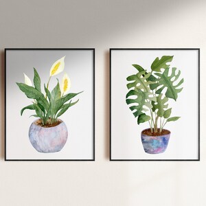 Op de afbeelding: Twee ingelijste aquarellen van potplanten. De afbeelding links toont een witte vredeslelie met groene bladeren in een lichtblauwe pot. De afbeelding rechts toont een groene monstera-plant in een paarse en blauwe pot.