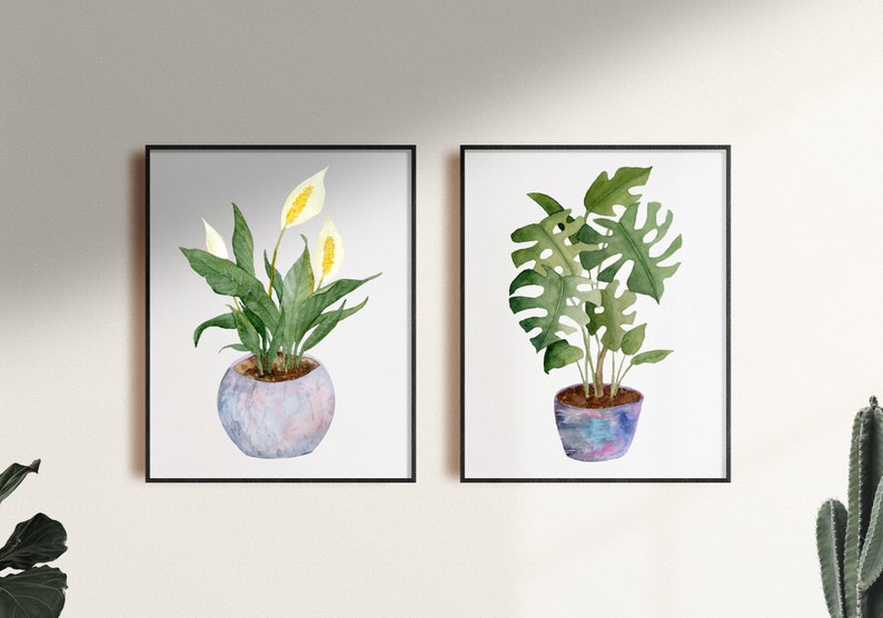 Op de afbeelding: Twee ingelijste aquarellen van potplanten. De afbeelding links toont een witte vredeslelie met groene bladeren in een lichtblauwe pot. De afbeelding rechts toont een groene monstera-plant in een paarse en blauwe pot.