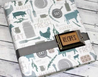 May include: A recipe binder with a farmhouse theme. The cover features a white background with gray and teal illustrations of kitchen utensils, farm animals, and text. A gray ribbon wraps around the binder, and a metal label reads "RECIPES".