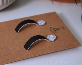 May include: A pair of black and white earrings with a marble design. The earrings are shaped like a curved line with a round marble-like charm at the end. The earrings are displayed on a brown card with the text "&" and "shop" printed on it.