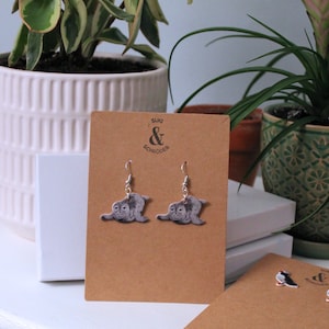 Puede incluir: Pendientes con forma de foca, grises y blancos, con ganchos plateados, presentados en una tarjeta marrón. La tarjeta tiene el texto "SUKI & SCHRODER" impreso. También se muestran otros pendientes.