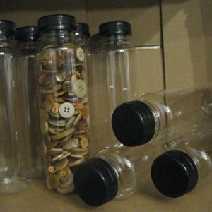 May include: Clear plastic bottles with black lids, some upright and some lying down. One bottle is filled with a variety of colorful buttons. The buttons are in shades of brown, beige, and white.