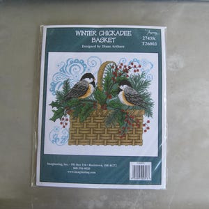 May include: A cross-stitch kit titled "Winter Chickadee Basket" featuring a detailed design of two chickadees perched on a basket filled with greenery and red berries. The kit includes the text "Designed by Diane Arthurs".