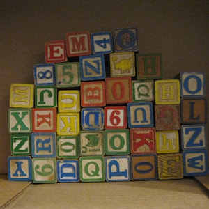 May include: A collection of vintage wooden alphabet blocks in various colors. Each block displays a letter or number, with some featuring small illustrations. The blocks are arranged in a pile, creating a playful and educational aesthetic.
