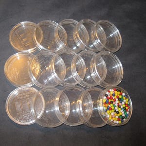 May include: A collection of clear, round plastic containers, some with lids, arranged on a dark surface. One container is filled with small, colorful spherical objects. The containers appear to be for storage or display.