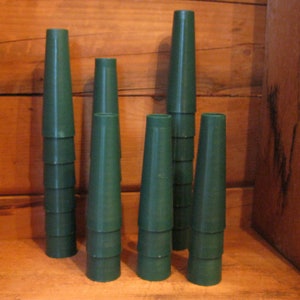 May include: A collection of six dark green, cone-shaped floral arranging tools. The tools are stacked and arranged in front of a wooden background. The tools are made of a smooth, solid material and have a flared top.