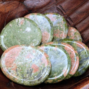 May include: A collection of polished unakite stones, each with a unique blend of green, pink, and orange hues. The stones are smooth and oval-shaped, displayed in a carved wooden bowl. The natural colors and patterns create a visually appealing arrangement.