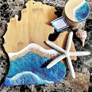 May include: A wooden cutting board shaped like the state of Michigan with a blue and white epoxy resin ocean wave design. The board is surrounded by a starfish, a seashell, and a small wooden box with a similar ocean wave design.