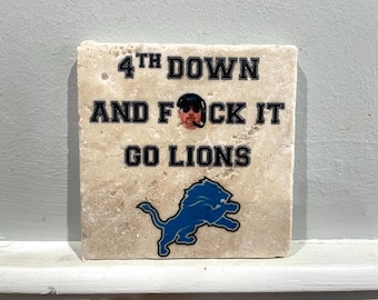 May include: A square, beige stone coaster with the text "4th DOWN AND F*CK IT GO LIONS" in black. A blue Detroit Lions logo is at the bottom. A small image of a person is in the center.