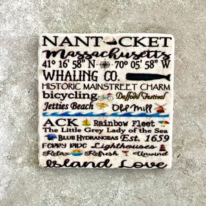 May include: A white stone coaster with black text and colorful illustrations. The text reads "NANTACKET Massachusetts 41° 16' 58" N 70° 05' 58" W WHALING CO. HISTORIC MAINSTREET CHARM bicycling Daffodil Festival Jetties Beach Old Mill ACK Rainbow Fleet The Little Grey Lady of the Sea BLUE HYDRANGEAS Est. 1659 FERRY RIDE Lighthouses Relax Refresh Unwind Island Love".