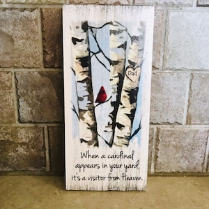 Puede incluir: Un letrero de madera con una imagen pintada de abedules y un cardenal rojo. El letrero tiene el texto "Pap&aacute;" en forma de coraz&oacute;n y el texto "Cuando un cardenal aparece en tu jard&iacute;n, es un visitante del cielo."