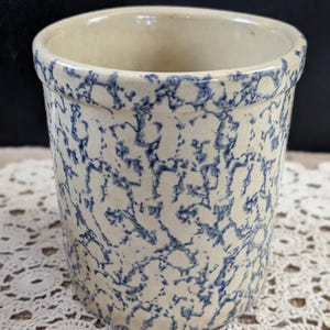 May include: A cylindrical stoneware crock with a cream-colored base and a blue spatterware design. The rim is slightly flared, and the interior is a solid cream color. This vintage kitchenware item is suitable for storage or display.